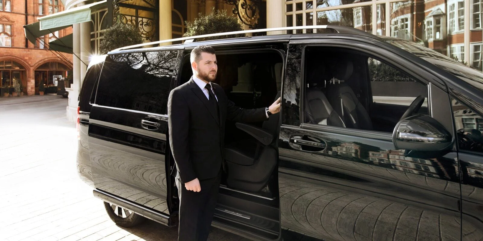 A professional chauffeur in a black suit standing next to a luxury Mercedes Business Van, ready for executive travel in London.